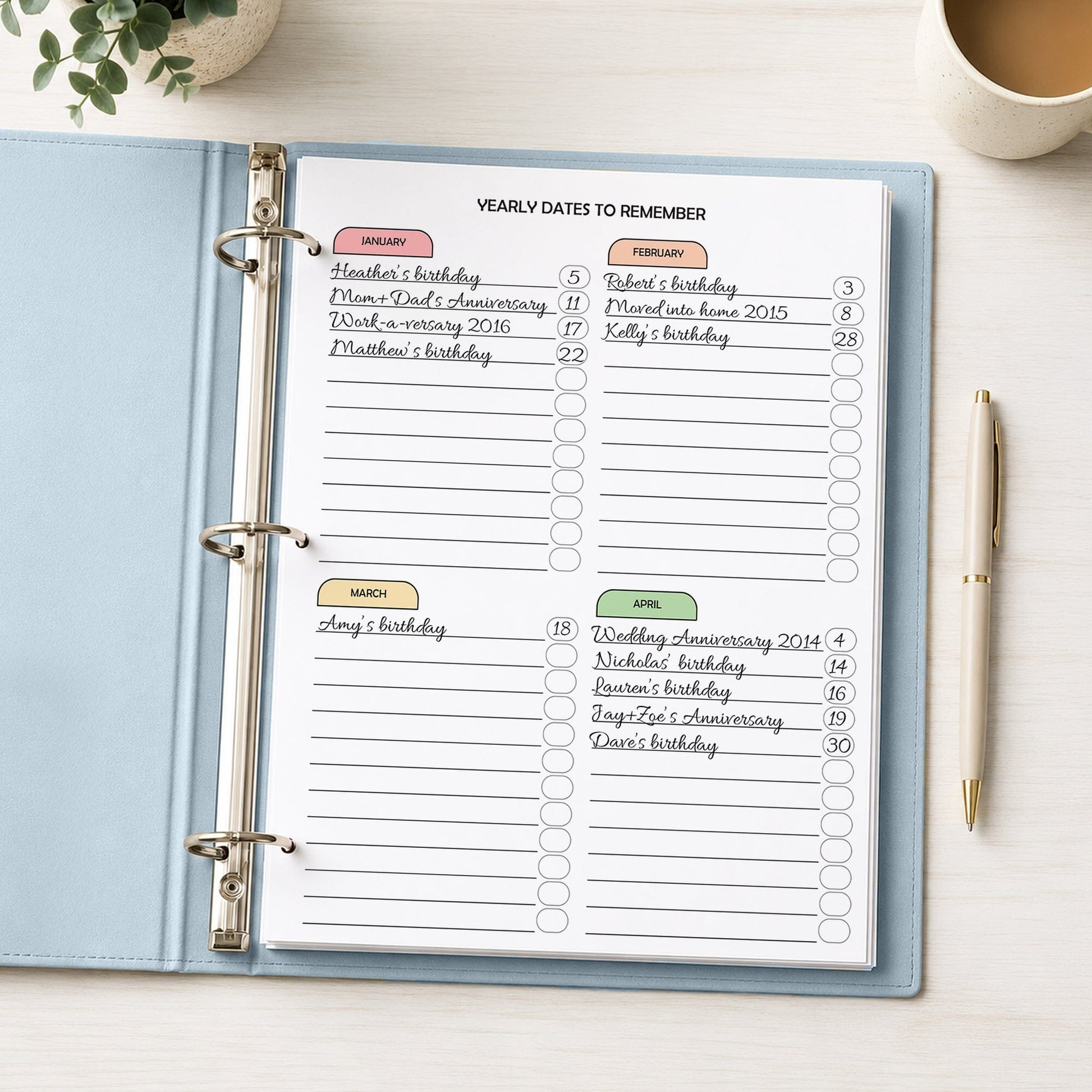 An open light blue ring binder planner showing printed monthly date reminder pages with handwritten entries, photographed on a white desk with a pen, plant, and coffee cup nearby.