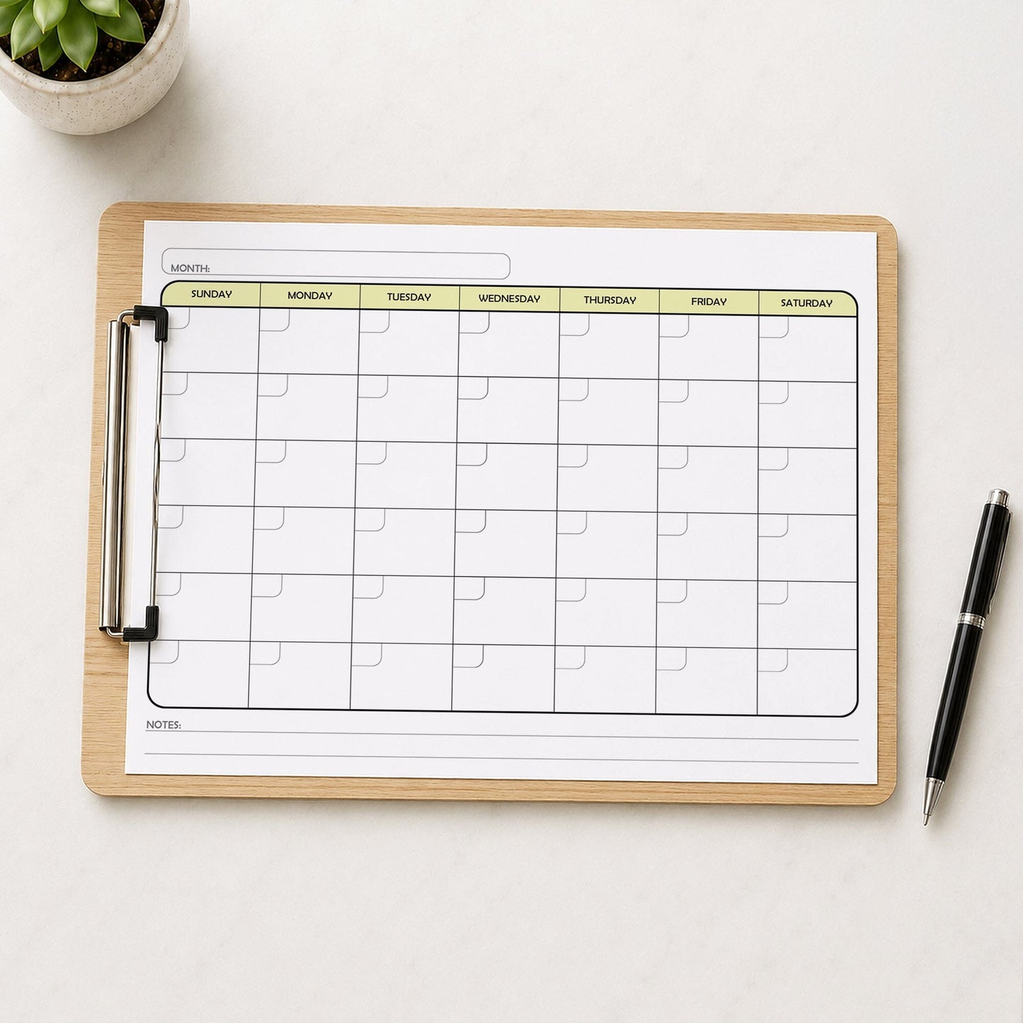 A printed Undated Monthly Calendar - Yellow Full-Page Planner (printable) page shown on a wooden clipboard on a table with a plant and a pen.