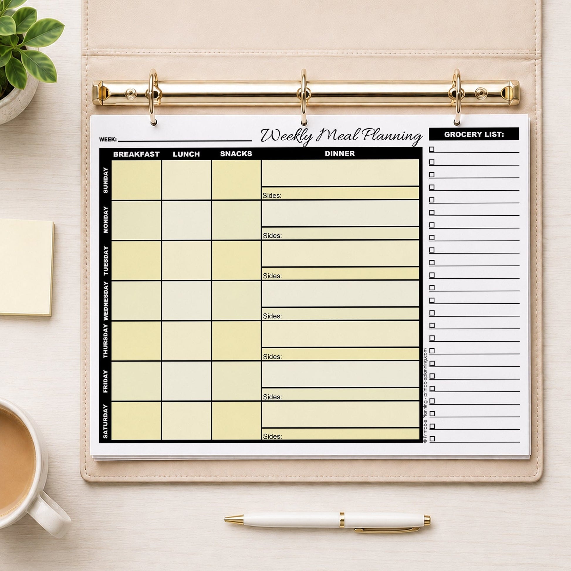 A printed Yellow Weekly Meal Planner with Grocery List (printable page) shown in a beige binder on a table with a plant, sticky notes, coffee, and a pen.