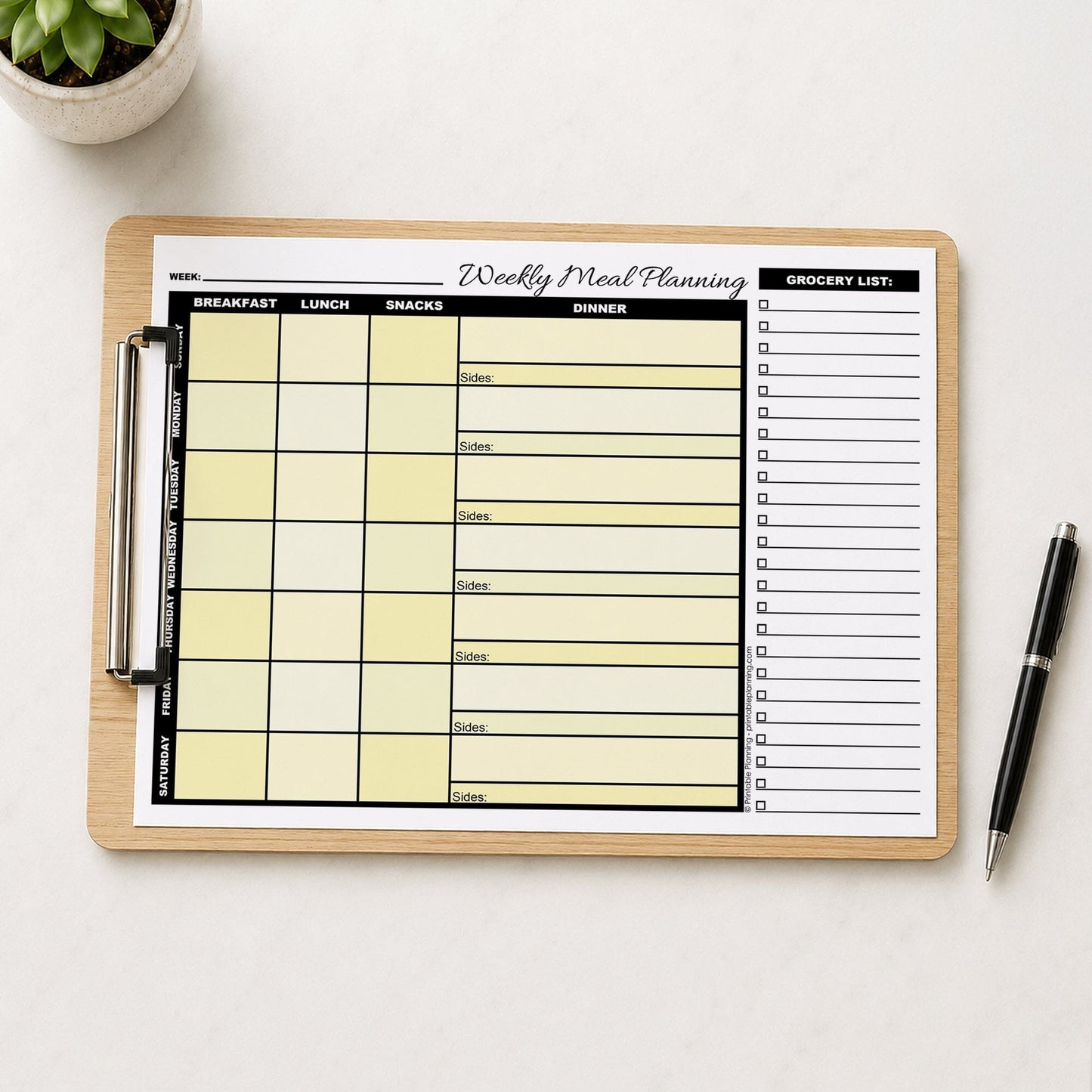 A printed Yellow Weekly Meal Planner with Grocery List (printable page) shown on a wooden clipboard on a table with a plant and a pen.